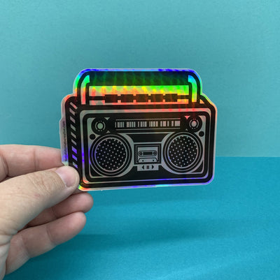 hand holding a holographic die cut sticker of a boom box illustration on a blue background