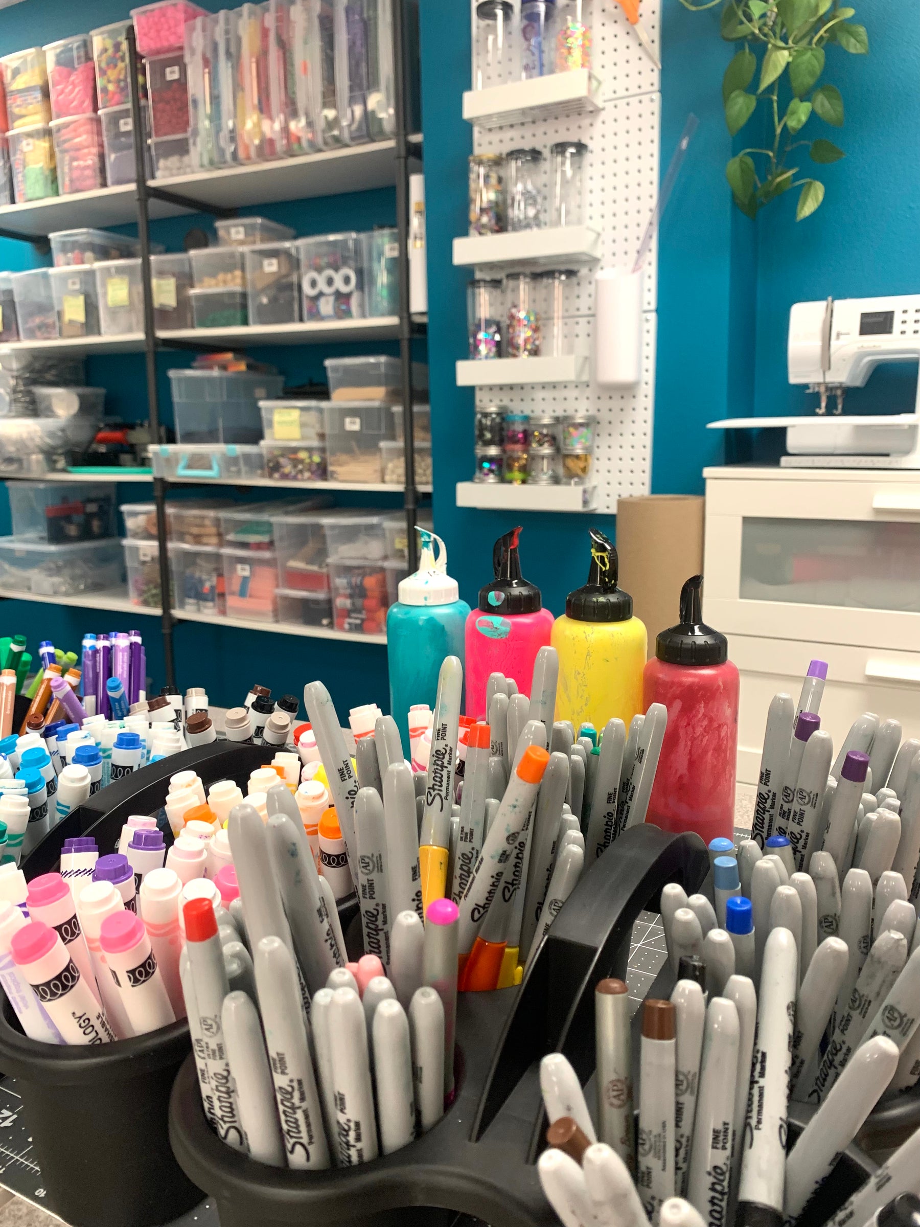 markers and sharpies in cups with paint bottles on a table. Shelves with storage for creative supplies are in the background