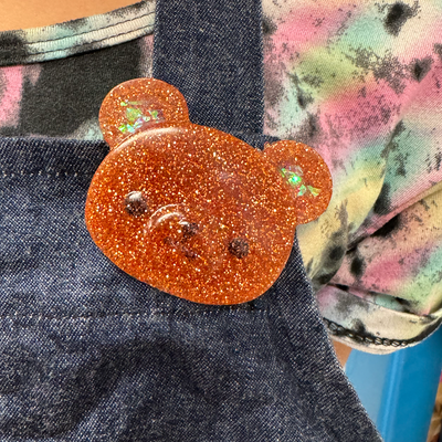 Glittery orange bear-shaped pin on a denim surface with a colorful background