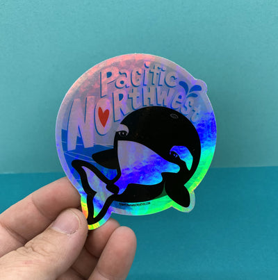 Hand holding holographic circle sticker of a whale and the words Pacific Northwest. Image is on a blue background